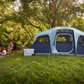 A large Coleman 10 person tent in two-tone blue in a grassy park with trees and a family sitting outside