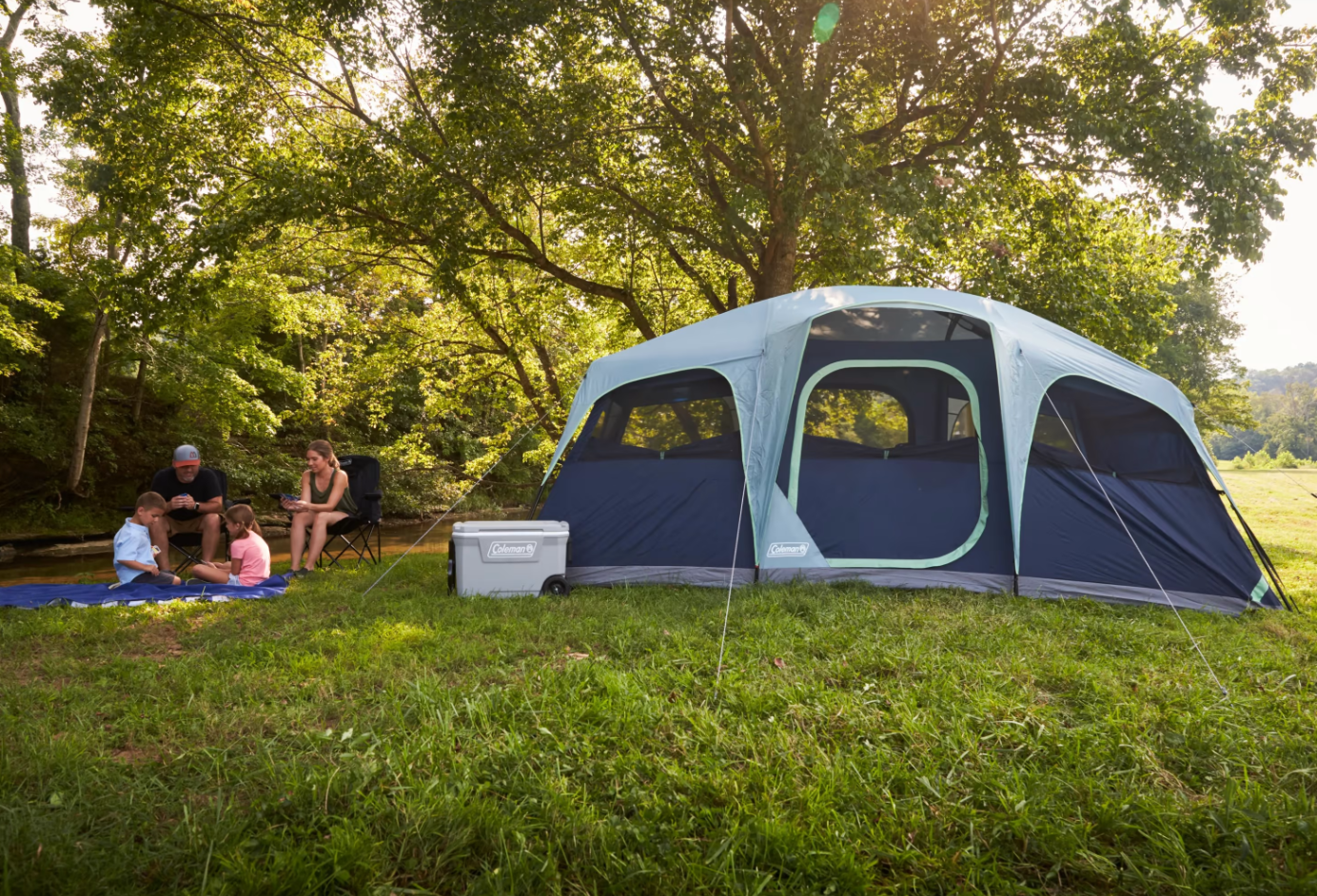 A large Coleman 10 person tent in two-tone blue in a grassy park with trees and a family sitting outside